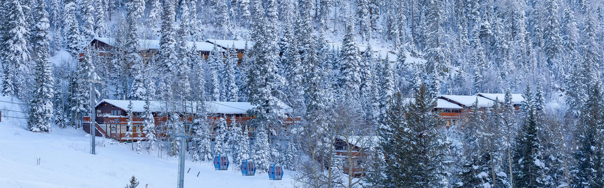 A snow covered wooden framed hotel sits perched above a ski run in the trees.