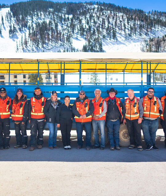 A group of people in orange safety vests pose in front of a blue shuttle car.