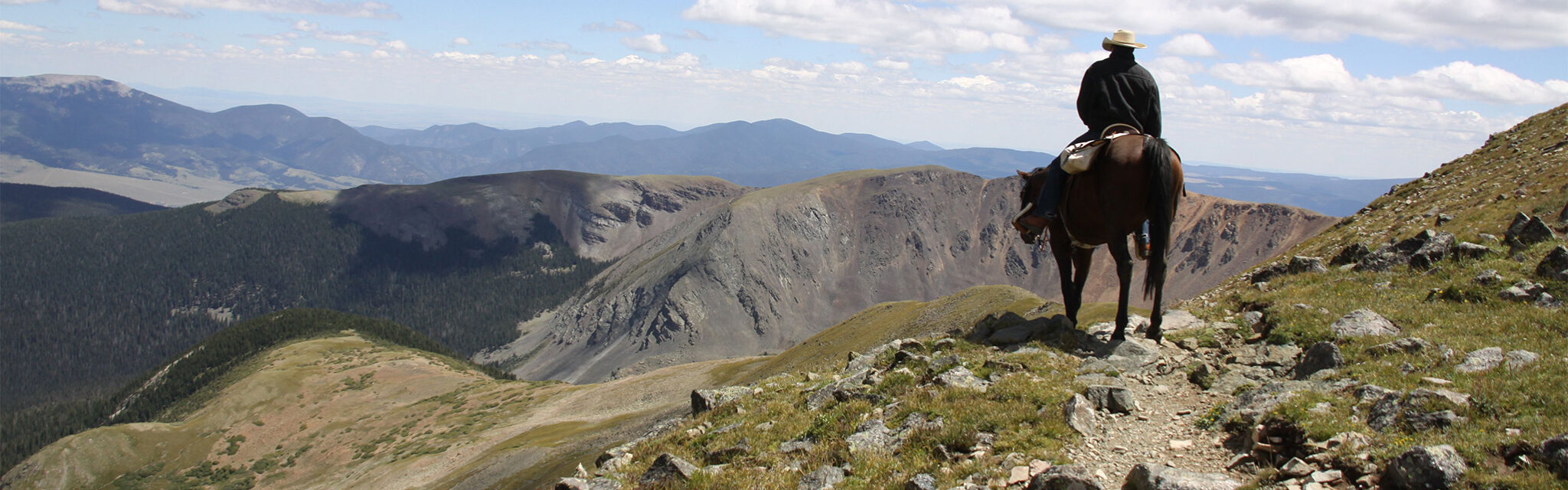 A lone horseback rider sits atop a mountain above treeline looking down at the valleys below.