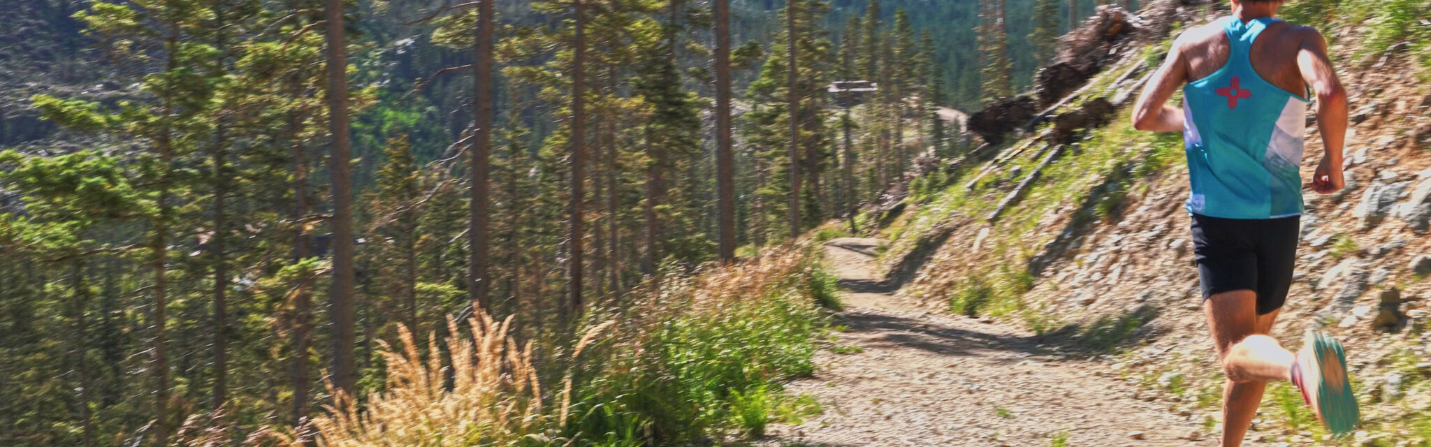 A man in a turquoise tank top runs down an open trail in the mountains.
