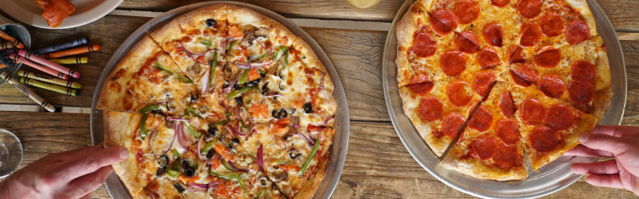 Two people's hands grab slices of pizza off a table with wings and crayons to the side.