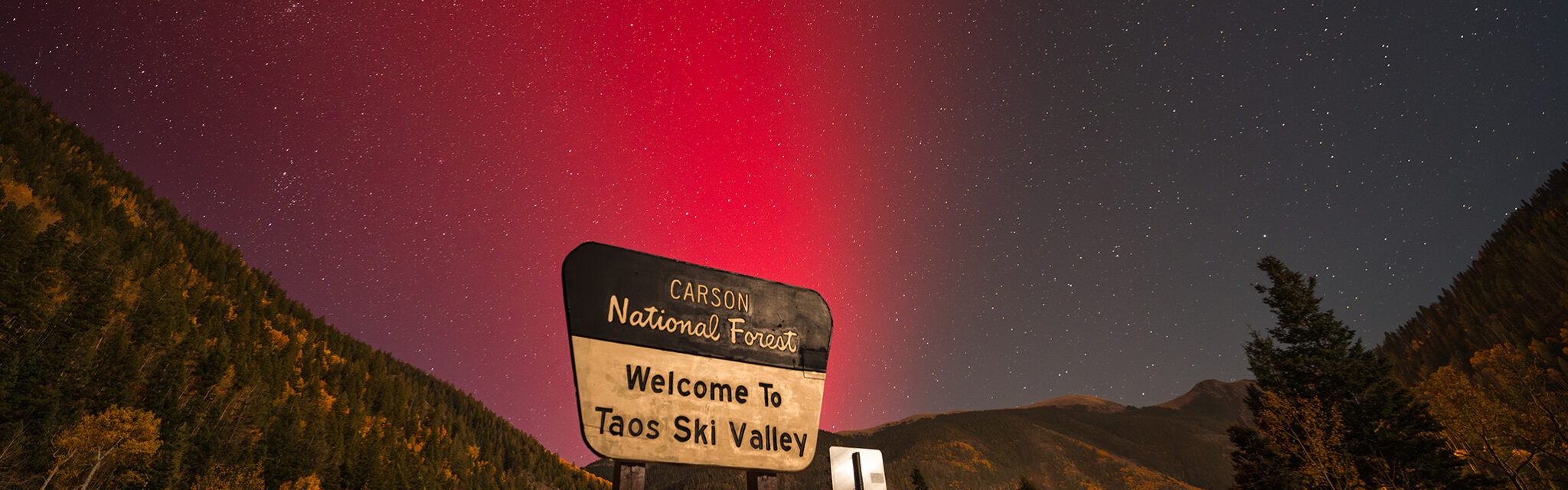 Magenta northen lights appear above a 'Welcome to Taos Ski Valley' sign.