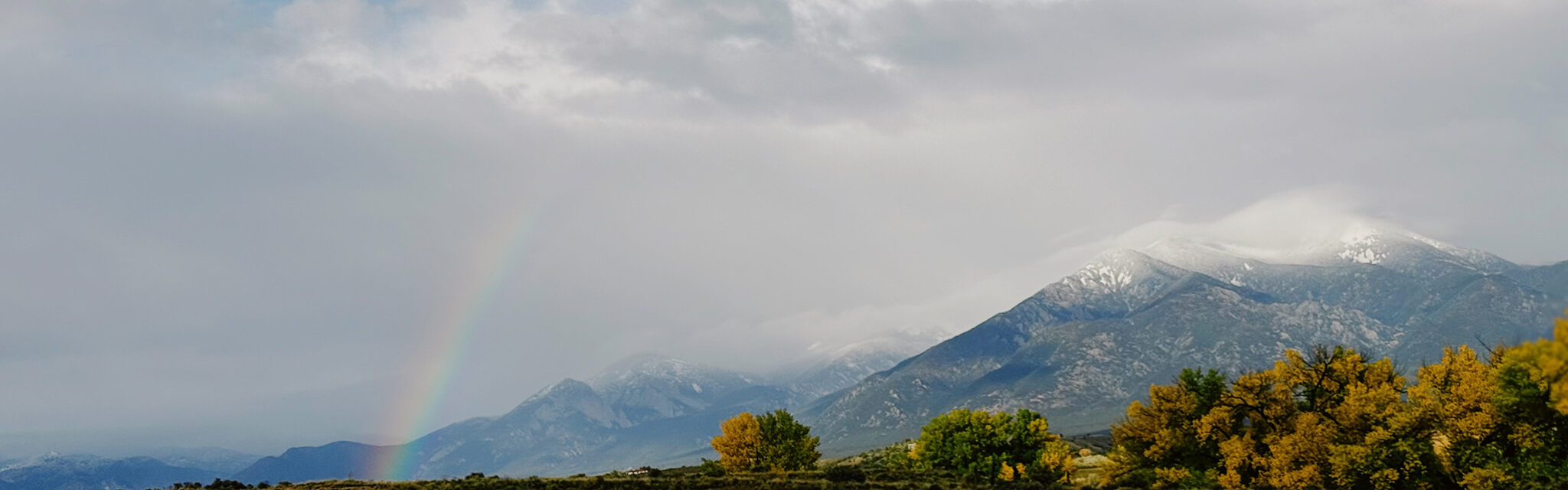 A rainbow reaches above a snow covered mountain.