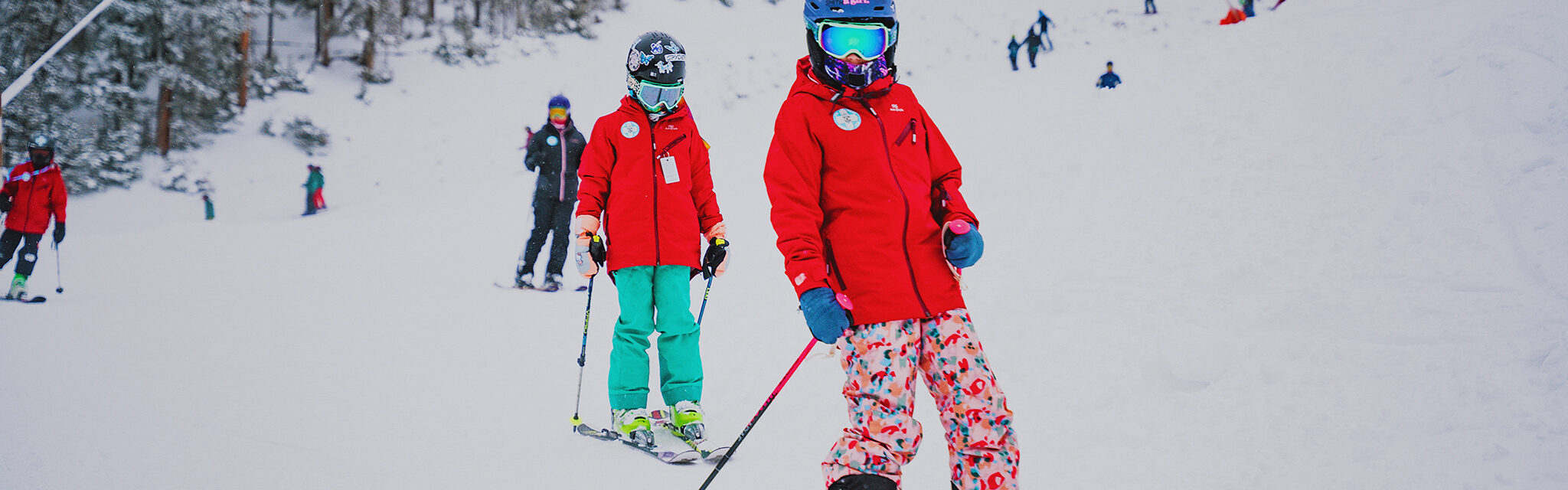 Two girls in red jackets and blue helmets ski past. A sticker on one helmet reads, "Ski like a girl"