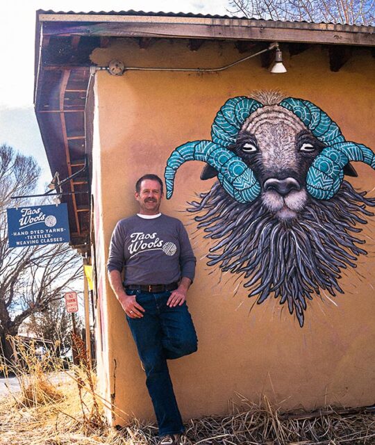 Taos Wools owner Joe standing next to the large sheep mural.