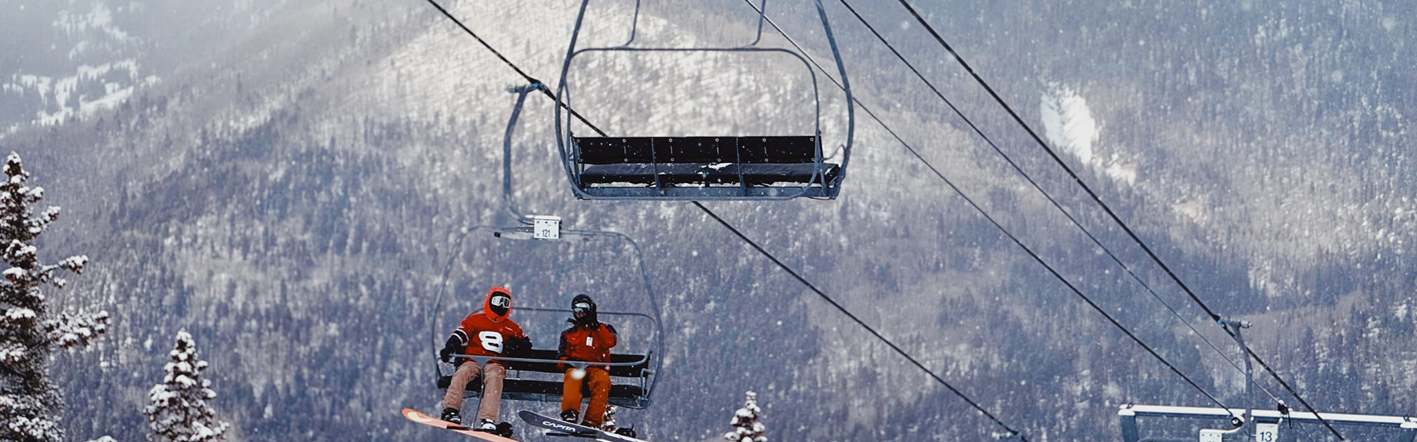 Two friends chat as they ride up a ski lift in the snow.