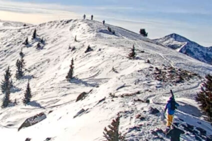 Two skiers hike a snowy path across a mountain ridge while carrying their skis.