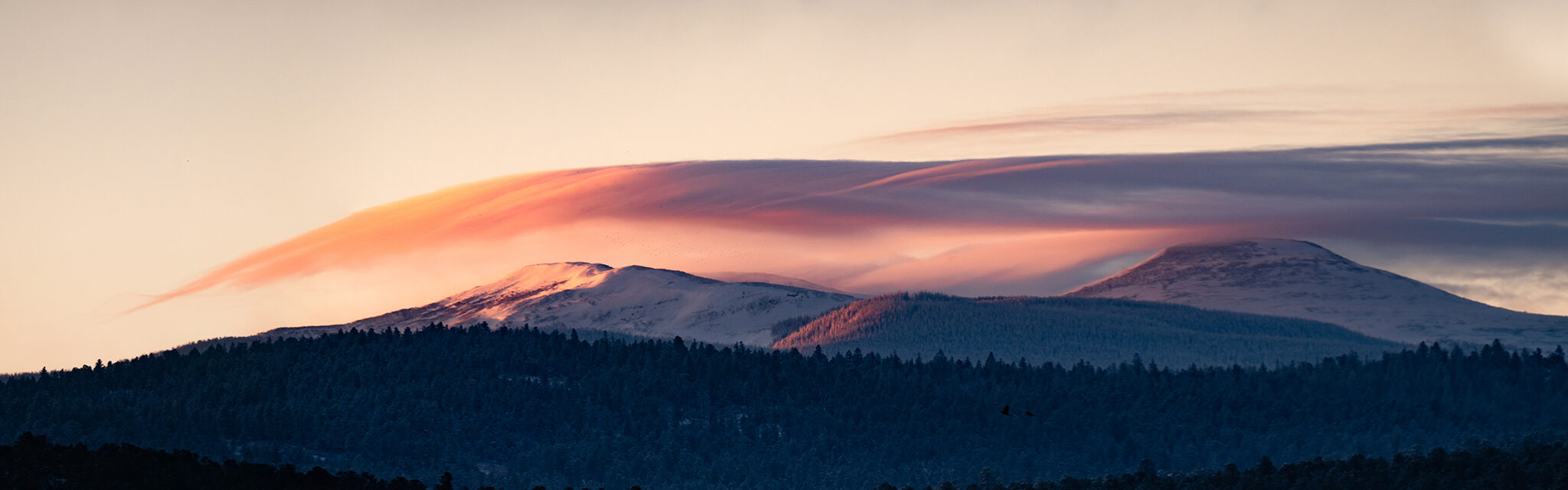 The light of sunrise illuminate a snowy mountains and smooth, wavelike clouds.