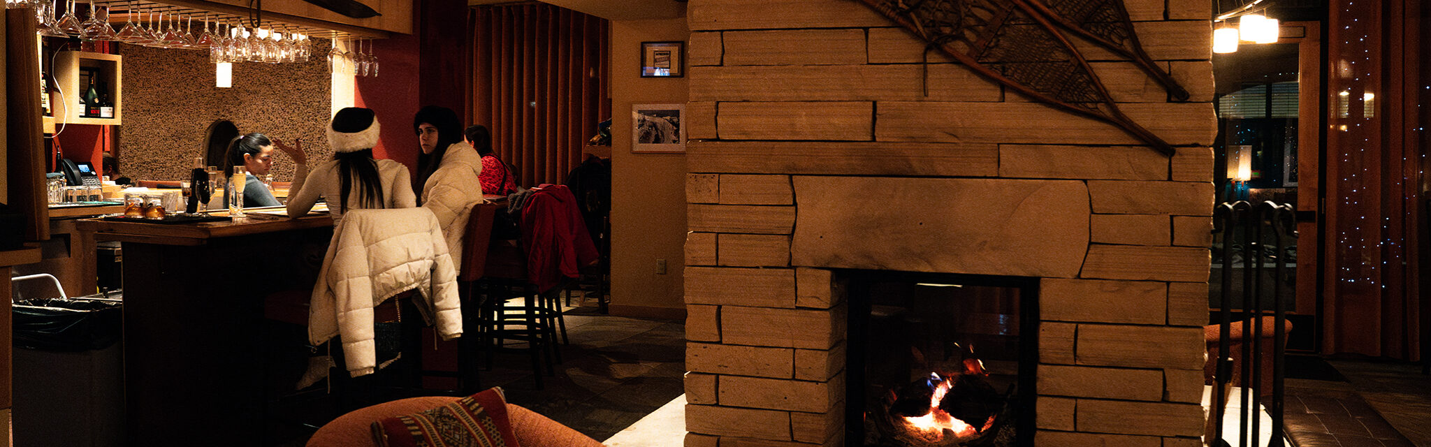 A dimly lit bar and lounge with a fireplace in the forefront and plush chairs.