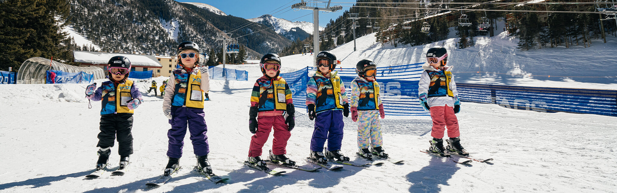 A group of kids wearing brightly colored snow gear stand in a line before a ski lesson.