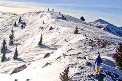 Two skiers hike a snowy path across a mountain ridge while carrying their skis.