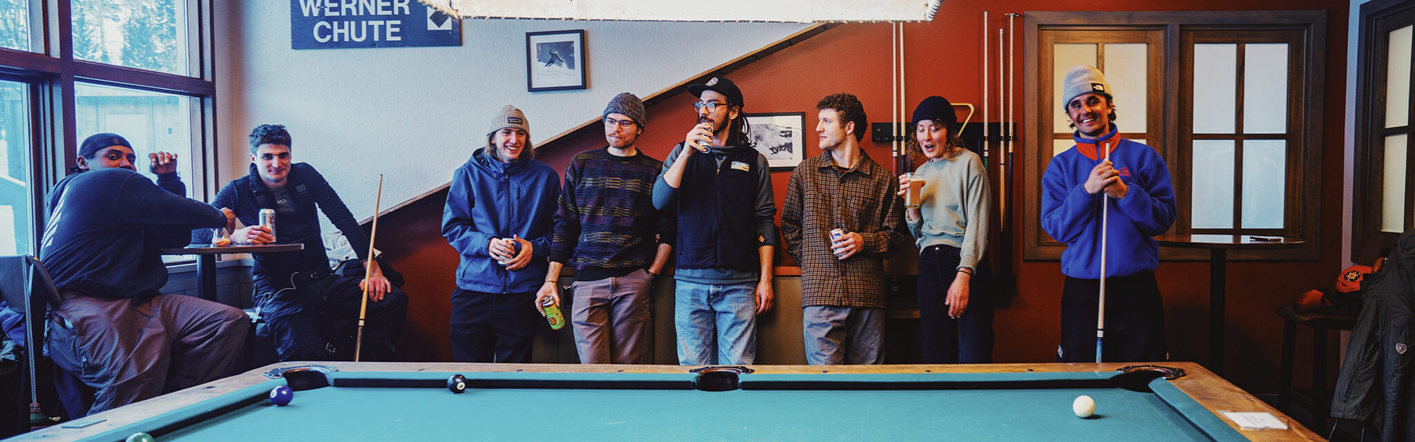 Friends stand in a line against in the wall in front of a pool table, chatting and laughing