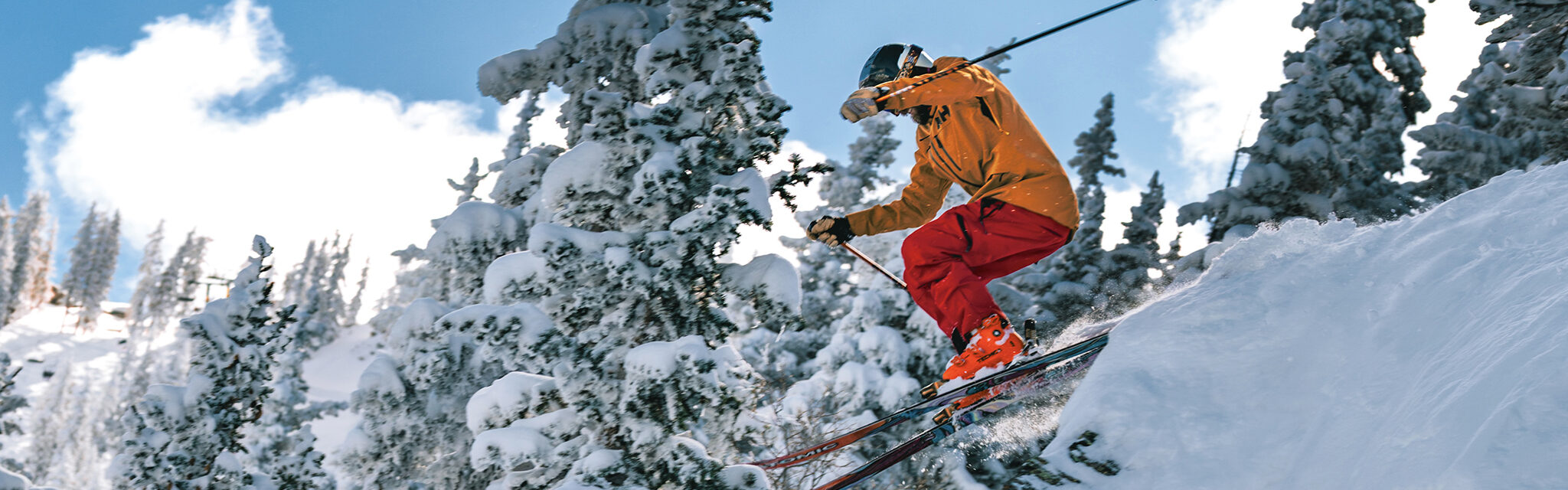 A skier in an orange jacket and red pants jumps off a snowy ledge.