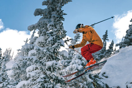 A skier in an orange jacket and red pants jumps off a snowy ledge.