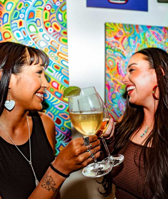 Two young women enjoying white wine with colorful art in the background.