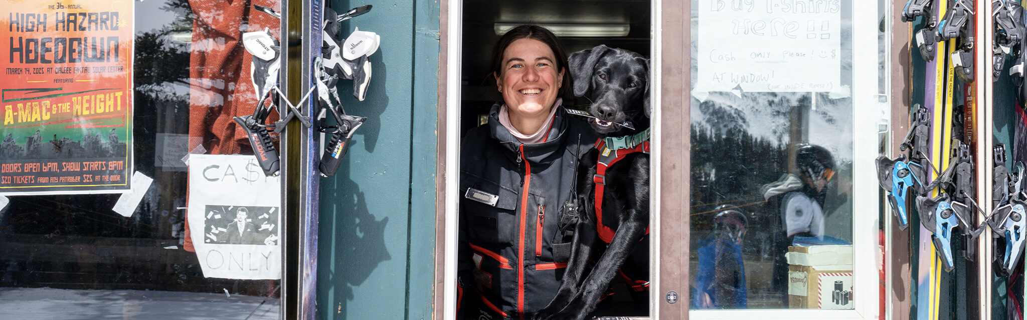 A woman and a small black labrador smile out an open window.