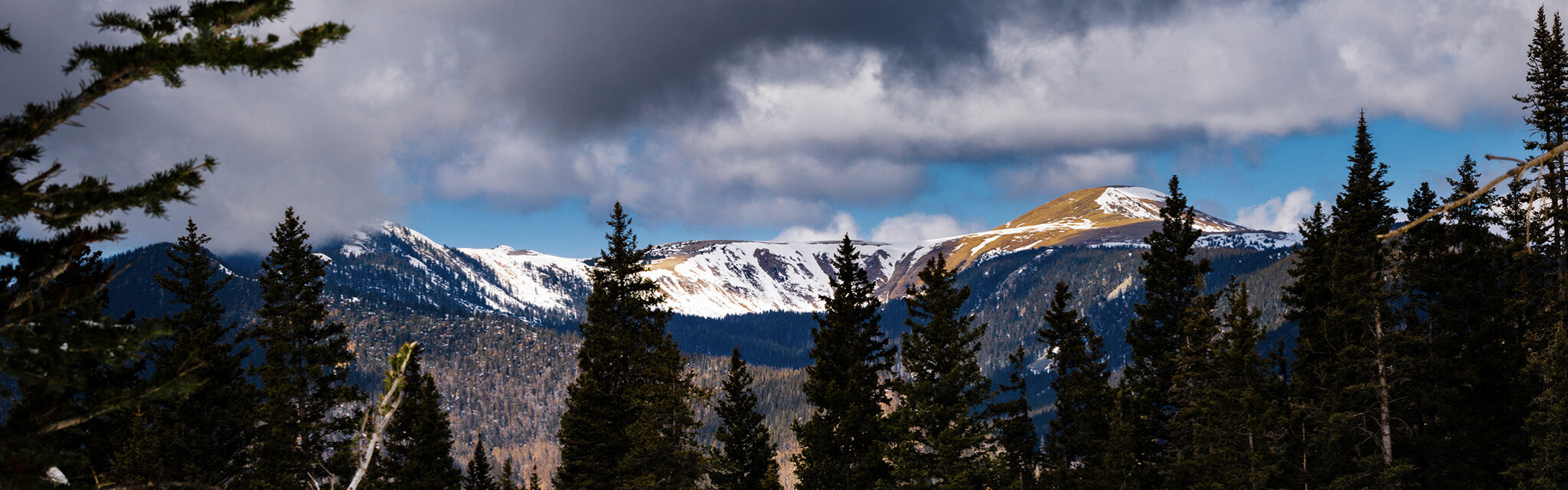 A partially snow covered mountain with ominous dark clouds above, but blue sky in the distance.