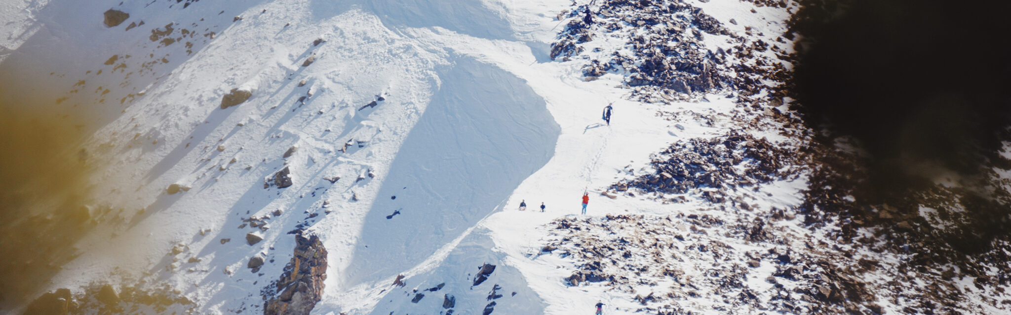 A distant view of brightly clad skiers and snowboarders hike up a ridgeline.