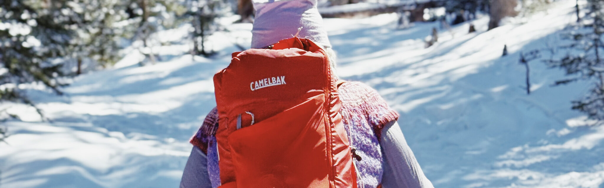 A woman wearing a pink hat and a red backpack walks away along a snowy trail.