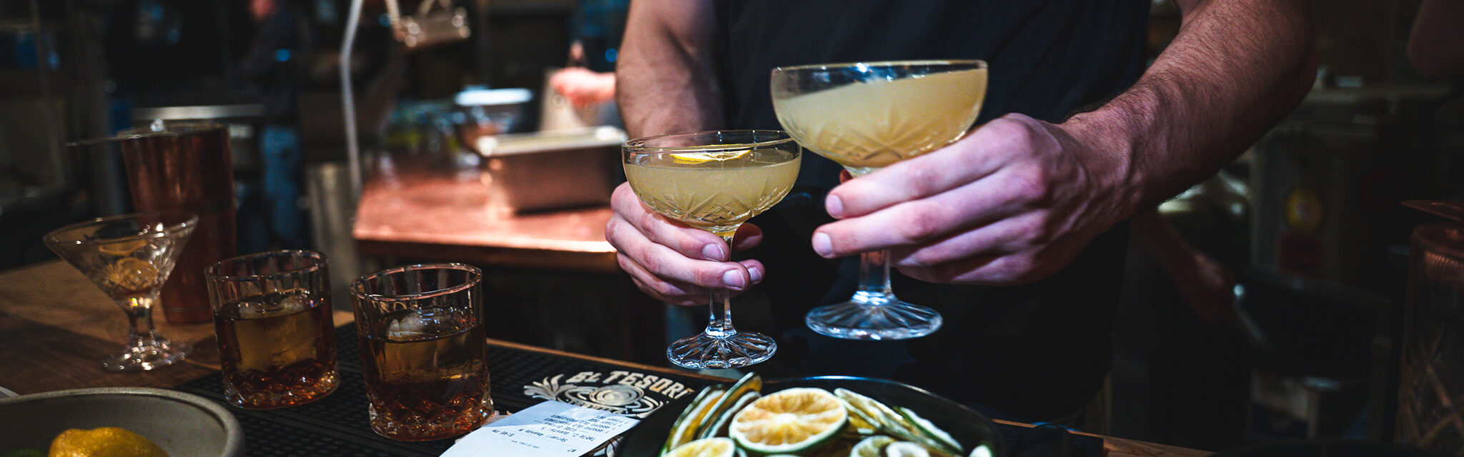 A bartender holds two margarita glasses, filled with cocktails while two whiskey drinks wait on the bar.