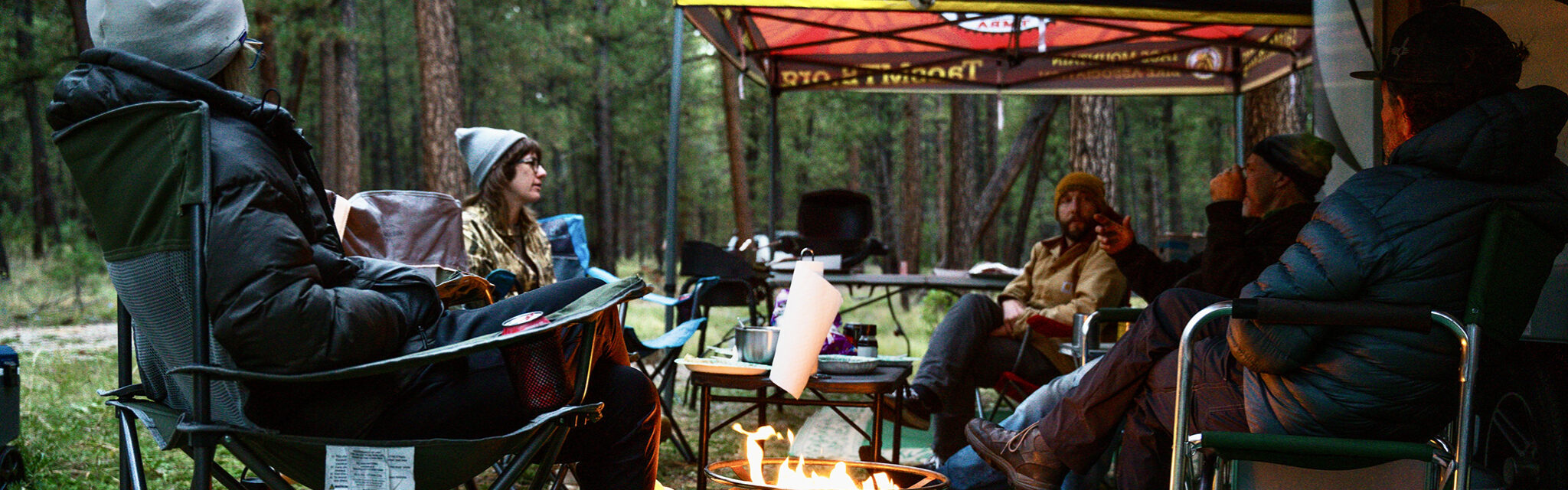 A group of people sit around a campfire in the woods under brightly colored awnings.