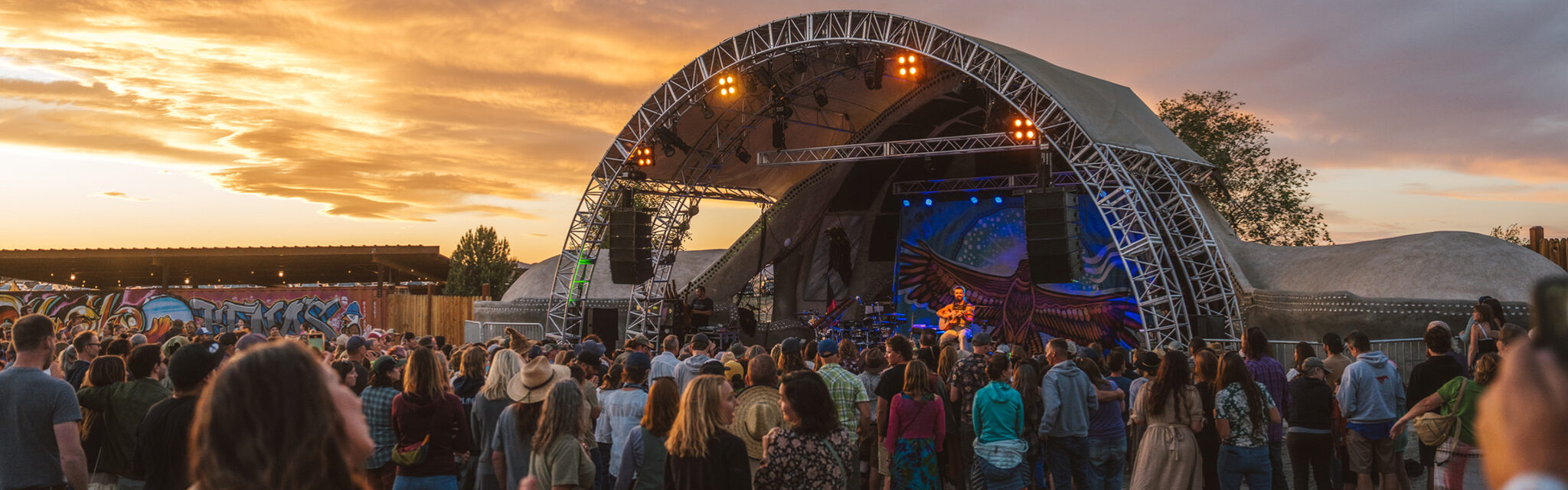 Sunset colors an outdoor concert with people dancing in front of an arched stage.