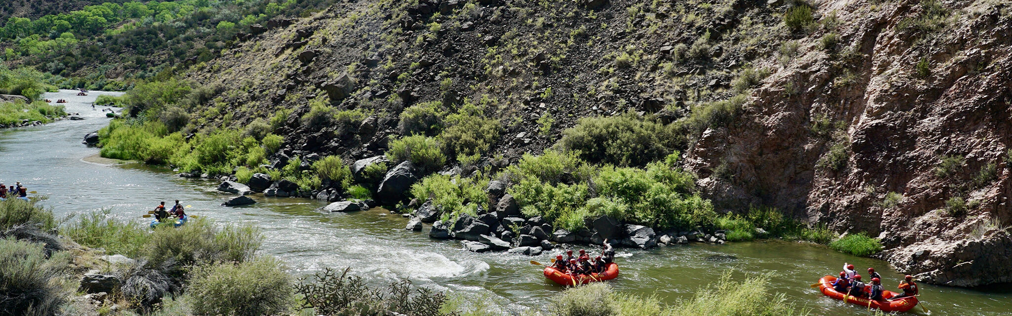 A group of river rafts make their way down a winding river in the desert.