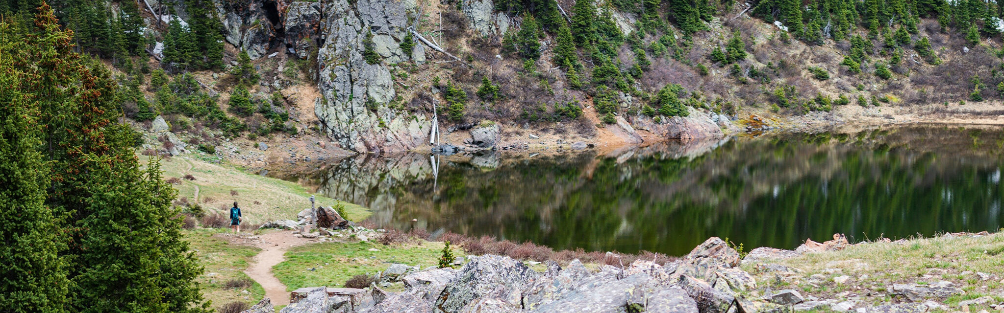 A lone hiker walks along the banks of a small lake surrounded by rocky slopes.