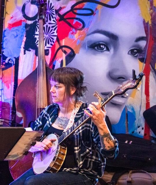 Woman playing a banjo with a large mural of a woman's head in the background.