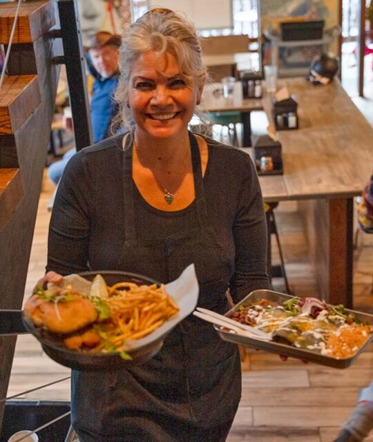 A smiling woman server holding plates of food at DALEEE TSV in Taos Ski Valley.