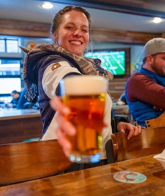 A young woman holding out a glass of beer towards the camera in a bar.