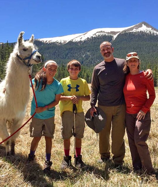 A family posing with a llama and snow on the mountains in the background with Wild Earth Llama Adventures.